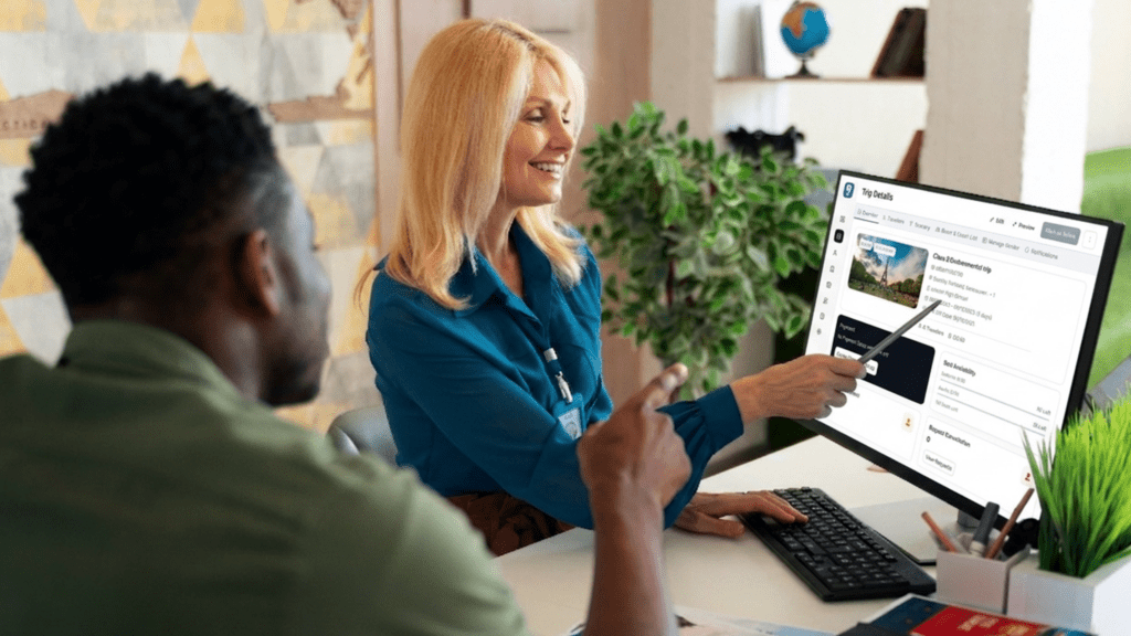 A smiling travel agent uses a stylus to point at "Trip Details" on a computer monitor running tour operator CRM software while consulting with a client.