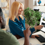 A smiling travel agent uses a stylus to point at "Trip Details" on a computer monitor running tour operator CRM software while consulting with a client.